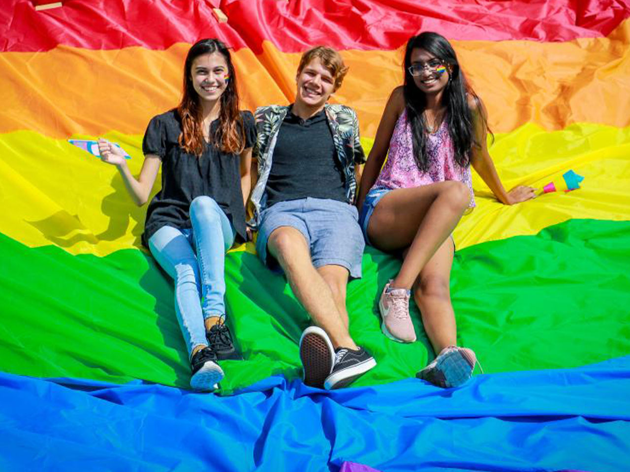 Three students seated on a pride flag out on the lawn