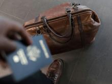 A hand, holding a passport, reaches from outside the frame toward a packed travel bag